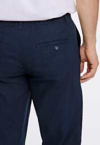 Pantaloni blu navy realizzati in tessuto leggero, con frontale piatto, vita elastica e tasca posteriore con bottone. La texture appare liscia.