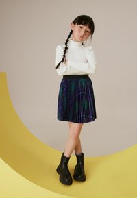 Schwarzer, plissierter Rock mit einem lila und grünen Muster, kombiniert mit einem weißen, gerippten Rollkragenpullover und schwarzen Stiefeletten, die auf einer gelben Kurve stehen.