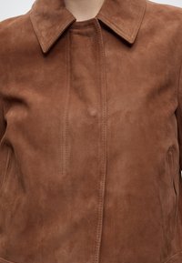 Veste en daim marron avec un col pointed et des détails de couture visibles, montrée du torse jusqu'à la zone du cou.