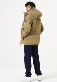 Veste isolée kaki avec capuche, dotée de poches zippées et d'un patch logo sur l'épaule, associée à un jean sombre et des baskets blanches.