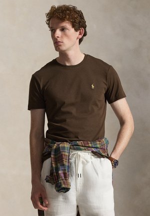 Młody mężczyzna z kręconymi włosami, ubrany w brązową koszulkę z krótkim rękawem, białe spodnie ze sznurkiem, koszulę w kratę zawiązaną wokół talii i zegarek na rękę.