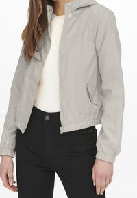 Světle šedá mikina na zip s kapucí, elastickými manžetami a lemem, se bočními kapsami, kombinovaná s bílým svetrem a černými džínami.