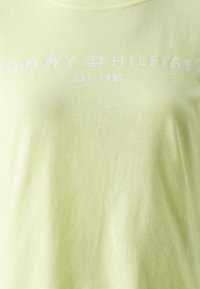 Tela amarilla claro con texto blanco "Tommy Hilfiger EST. 1985" y un pequeño logotipo rectangular en el centro.