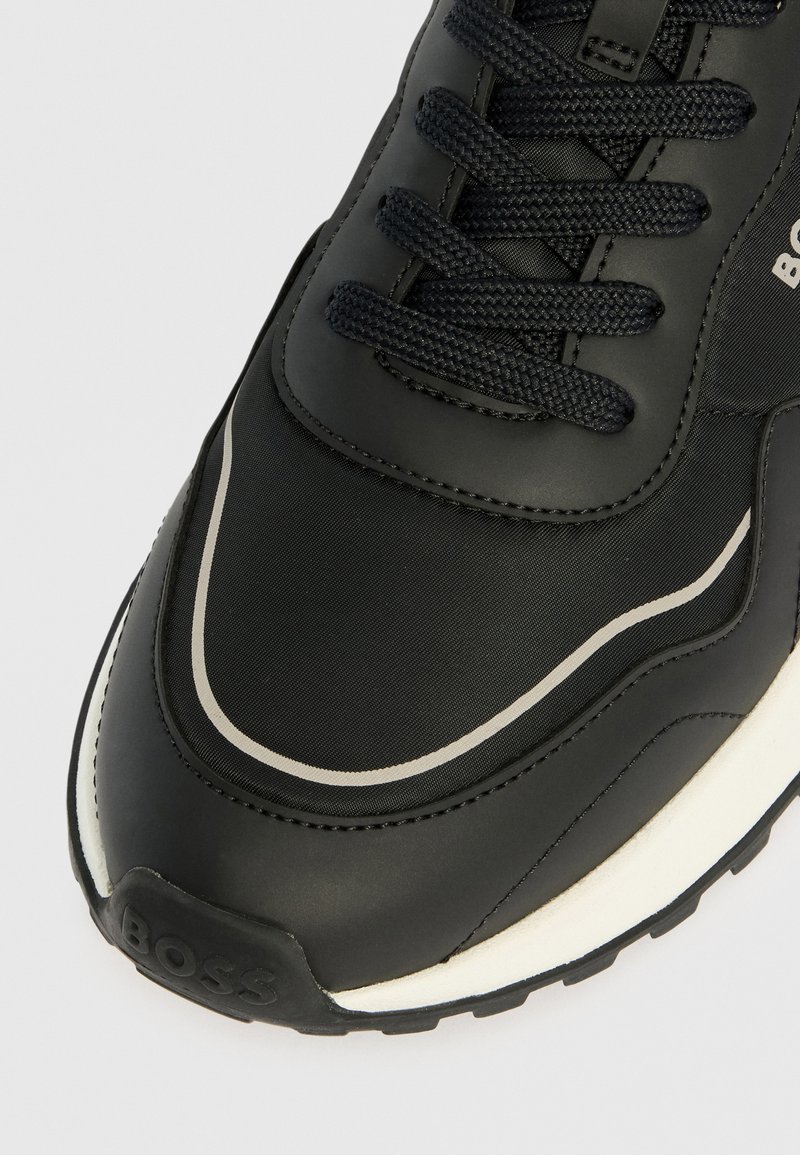 Baskets noires avec lacets noirs, semelle blanche, dessus en tissu et cuir, et logo "BOSS" en relief sur l'embout avant.