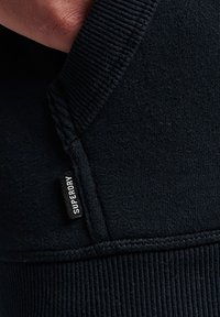 Felpa nera con polsini e orlo a costine, tessuto strutturato e una piccola etichetta in tessuto con la scritta "SUPERDRY" sulla cucitura laterale.