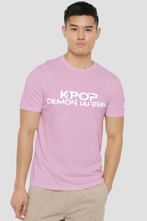 Junger Mann trägt ein pinkes T-Shirt mit dem Text „K-POP DEMON HUNTERS“ und steht vor einem schlichten hellgrauen Hintergrund.