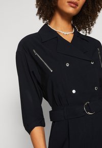 Femme portant une veste noire ceinturée avec des boutons argentés, des poches poitrine zippées, des manches trois-quarts et un collier de perles.