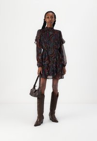 Vestido escuro com padrão paisley, com mangas em babados e gola alta, combinado com botas de cowboy castanhas e uma pequena mala de ombro.