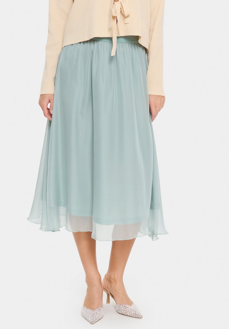 Gonna midi in turchese chiaro con un design morbido e fluente, stile a strati e vita elastica. Indossata con un maglione beige a coste e tacchi decorati.
