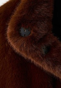 Tessuto in pelliccia sintetica marrone con una finitura morbida e testurizzata. Bottoni neri visibili sul colletto, che aggiungono dettagli funzionali e di design.