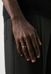 Zilveren bandring om een hand met een donkere huidtint, met een gladde oppervlakte en een eenvoudig geometrisch ontwerp. De hand rust op donkere stof.