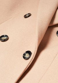 Abrigo cruzado en tela rosa suave, con botones negros de diseño circular y textura. Líneas limpias y forma entallada.