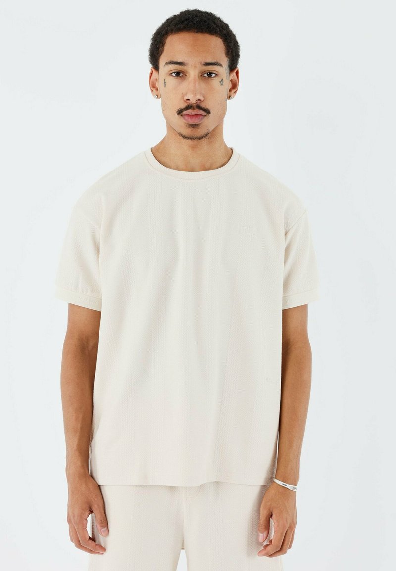 Camiseta de color crema, de corte holgado, con mangas cortas, tela texturizada, cuello redondo y un sutil detalle de logo en la parte frontal.