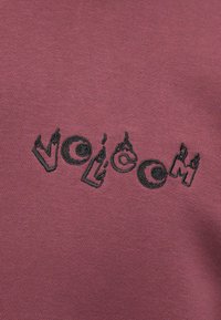 Sweatshirt bordeaux avec logo "VOLCOM" brodé en noir, présentant des accents de flamme sur les lettres, texture douce et coupe décontractée.
