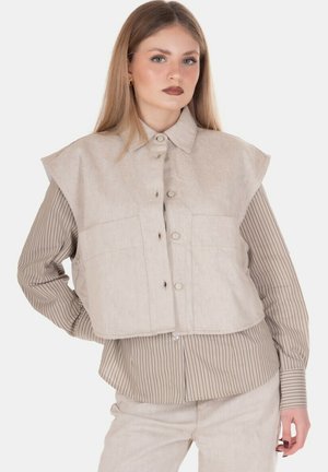 Giovane donna con lunghi capelli biondi che indossa un gilet beige senza maniche con bottoni sopra una camicia marrone a righe a maniche lunghe e pantaloni coordinati.