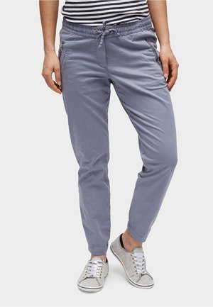 Personne portant un pantalon bleu clair à cordon avec poches zippées et des baskets grises décontractées, debout les jambes croisées.