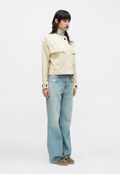 Άτομο που στέκεται, φορώντας μπεζ cropped μπουφάν, πουλόβερ με ριγέ ζιβάγκο, φαρδιά γαλάζια τζιν παντελόνια και ελιά πράσινα αθλητικά παπούτσια σε λευκό φόντο.