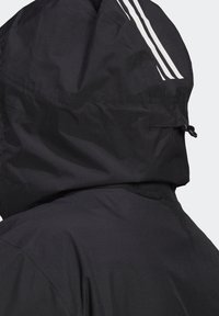 Veste noire avec capuche, texture lisse et trois bandes blanches sur le côté. Le tissu semble léger et résistant à l'eau.