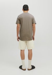 Camiseta marrón de manga corta, pantalones cortos crema, calcetines blancos y sandalias negras; observando un diseño simple con un ajuste relajado.