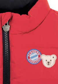 Red padded jacket featuring a black zipper, bear-shaped zipper pull, FC Bayern München emblem, and a fluffy white bear patch.