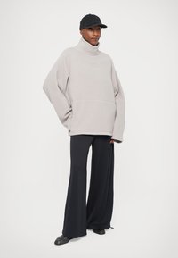 Jasnoszary polarowy sweter z wysokim kołnierzem i przednią kieszenią, zestawiony z czarnymi szerokimi spodniami o fakturze oraz czarnymi butami na slip-on.