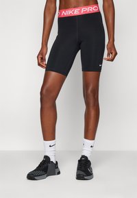 Fekete Nike Pro short, rózsaszín derékkal, térd hosszig érő fazon, sima textúrával, és fehér Nike logóval az alsó lábszáron. Fekete cipővel viselve.