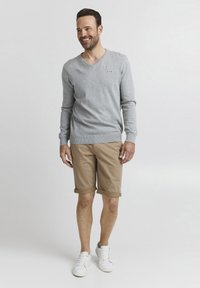 Grå genser med v-hals, beige rullede shorts, og hvite sneakers. Genseren har små logo detaljer. Modellen står og smiler mot en ensfarget bakgrunn.