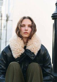 Chaqueta de cuero negra con un gran y esponjoso cuello color crema, llevada sobre un suéter beige, combinada con pantalones verde oliva; sentado contra un fondo claro.
