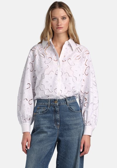 Frau trägt weiße, geblümte Bluse mit Knöpfen und Lochstickerei, gepuffte Ärmel und hoch taillierte blaue Jeans, steht vor einfachem Hintergrund.