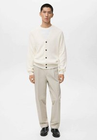 Cárdigan blanco de canalé con botones marrones, combinado con pantalones beige claros y zapatos negros, presentado sobre un fondo neutro.