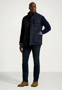 Moški v mornarsko modrem podložnem telovniku, oblečenem čez temno pulover, temne kavbojke in svetle usnjene čevlje, stoji z eno roko v žepu telovnika.