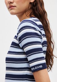 Ριμπ πλεκτό τοπ με οριζόντιες μπλε και λευκές ρίγες, κοντά μανίκια και ετικέτα με λογότυπο στη μανσέτα. Το ύφασμα φαίνεται μαλακό και εφαρμοστό.