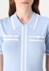 Ljust blå stickad polo skjorta med en spetsig krage, två vita randiga fickdetaljer och vertikala vita ränder fram.