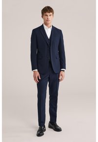 Młody mężczyzna stoi w granatowym trzyczęściowym garniturze, białej koszuli i czarnych eleganckich butach na jednolitym beżowym tle.