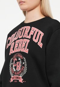 Zwarte sweatshirt met opvallende roze tekst "COLOURFUL REBEL," met een cirkelvormig logo en laurierontwerp aan de onderkant.