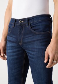 Mörkblå denimjeans med en åtsittande design, som har fem fickor, kontrasterande orange sömmar och en rund svart knapp i midjan.