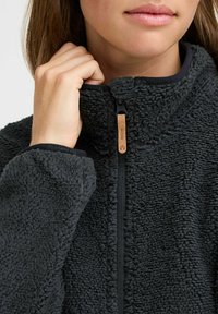 Tumehall fleece-jakk, millel on kõrge krae, tekstuuriga pind, eesmine lukku ja puidust tõmblukk. Varrukate ääred on musta ääremisega.