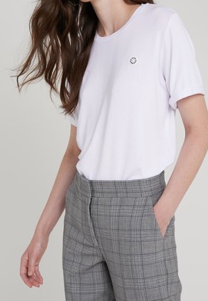 Woman wearing a plain white short-sleeve shirt with a small circular logo and gray plaid high-waisted pants with one hand in pocket.