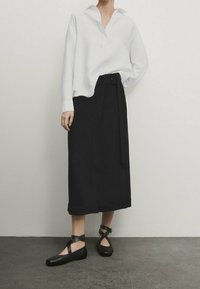 Blusa bianca a maniche lunghe con scollo a V, abbinata a una gonna nera a vita alta con un fiocco laterale. Scarpe flat nere con fasce incrociate.