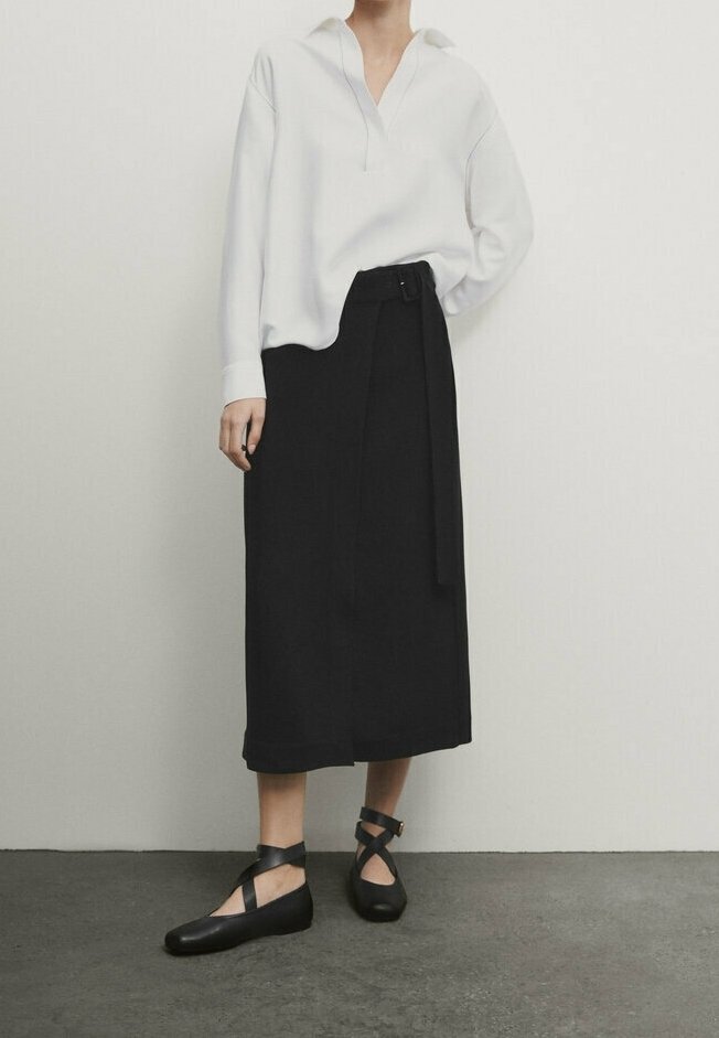 Blusa bianca a maniche lunghe con scollo a V, abbinata a una gonna nera a vita alta con un fiocco laterale. Scarpe flat nere con fasce incrociate.