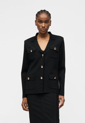 Γυναίκα που φοράει μαύρο σακάκι με χρυσά κουμπιά και τέσσερις μπροστινές τσέπες, σε συνδυασμό με μια μαύρη φούστα που ταιριάζει, σε λευκό φόντο.