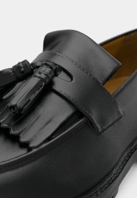 Gros plan sur un mocassin en cuir noir avec pompon et franges sur le cou-de-pied, montrant une texture lisse et des coutures.