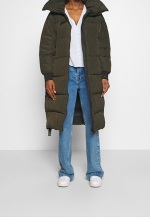 Person trägt einen olivgrünen langen Daunenmantel über einem weißen Hemd mit Knöpfen, blauen Jeans und weißen Turnschuhen und steht vor einem schlichten Hintergrund.