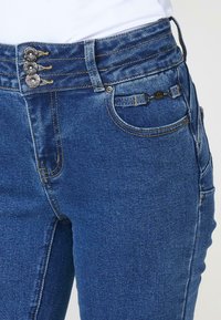 Vaqueros de tiro alto en azul oscuro con tres botones en la parte superior, costuras en contraste y bolsillos frontales con herrajes decorativos.