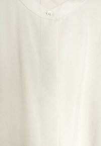 Lysebeige skjorte med V-udskæring, enkelt knaplukning og glat stoftekstur. Har lodrette syninger ned foran.