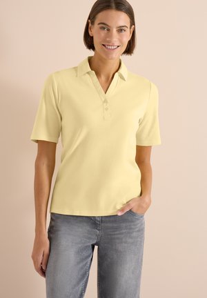 Kvinde smilende iført en sart gul kortærmet poloshirt og grå jeans, stående med den ene hånd i lommen mod en beige baggrund.