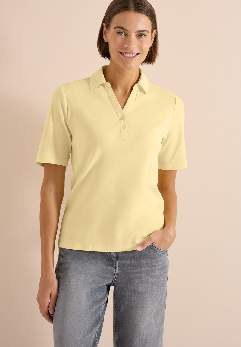 Femme souriante portant un polo jaune pâle à manches courtes et un jean gris, debout avec une main dans sa poche devant un fond beige.