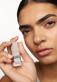 Transparante fles met zilveren dop, gelabeld "le mini macaron gel shine top coat." Close-up van hand en gezicht met een neutrale achtergrond.