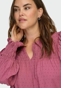 Kobieta o falujących brązowych włosach, nosząca srebrne kolczyki w kształcie kół i teksturowaną bluzkę w kolorze mauve z marszczonym dekoltem i bufkami, dotyka szyi.