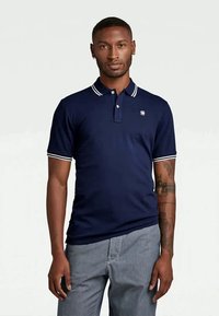 Muž v námorníckom polo tričku s bielymi pruhmi na golieri a rukávoch, a svetlo šedých pruhovaných nohaviciach, stojí na bielom pozadí.